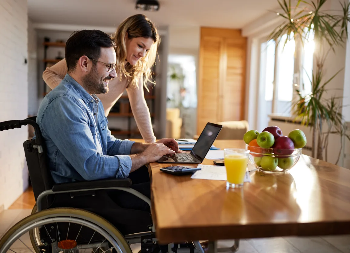 man in wheelchair and woman smiling at laptop during the day in dinning room