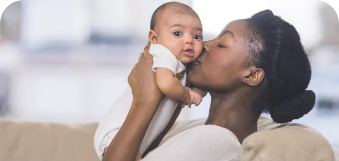 Mother holding baby near face kissing gently while smiling as the baby looks at the camera