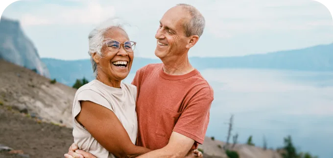 Elderly couple holding each other smiling near a lake with mountains in the background