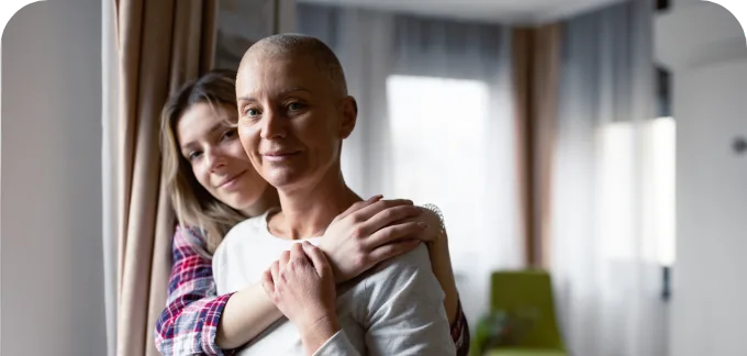 A smiling woman with a shaved head stands by a window, embraced by a younger woman in a plaid shirt. Their warm connection reflects the comfort found in oncology support and education, helping them navigate cancer care and emotional challenges together.