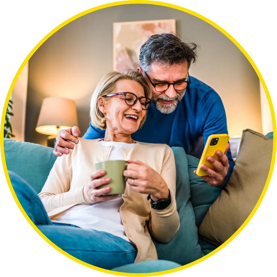Smiling older couple sitting on a
									couch at home. The woman is holding a mug
									while the man, with his arm around her,
									shows something on a yellow smartphone. They
									both appear happy and relaxed.