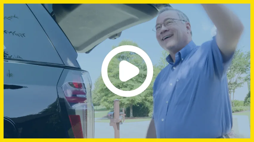 smiling older man lifting the back
										hatch of an suv in a sunny neighborhood
										driveway with play button overlay - links to video