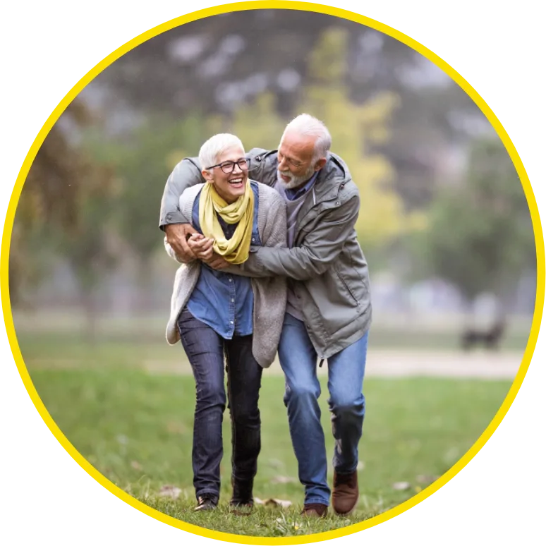 An elderly couple joyfully walks through a park, smiling and hugging. The scene conveys warmth and happiness, with a blurred, green background.