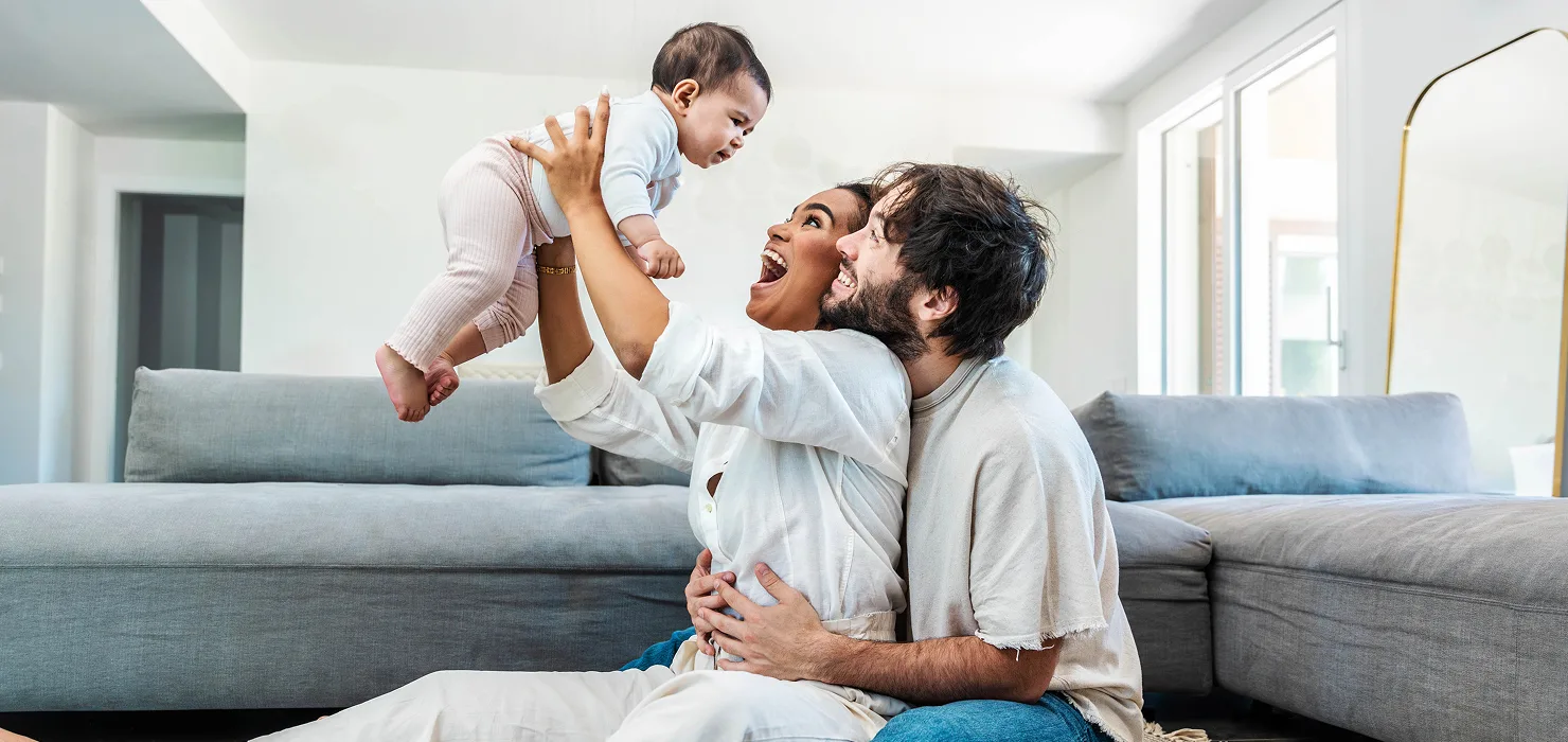 young couple at home sitting on the floor of the living room in front of the couch with the mother holding her baby up & both smiling at the child