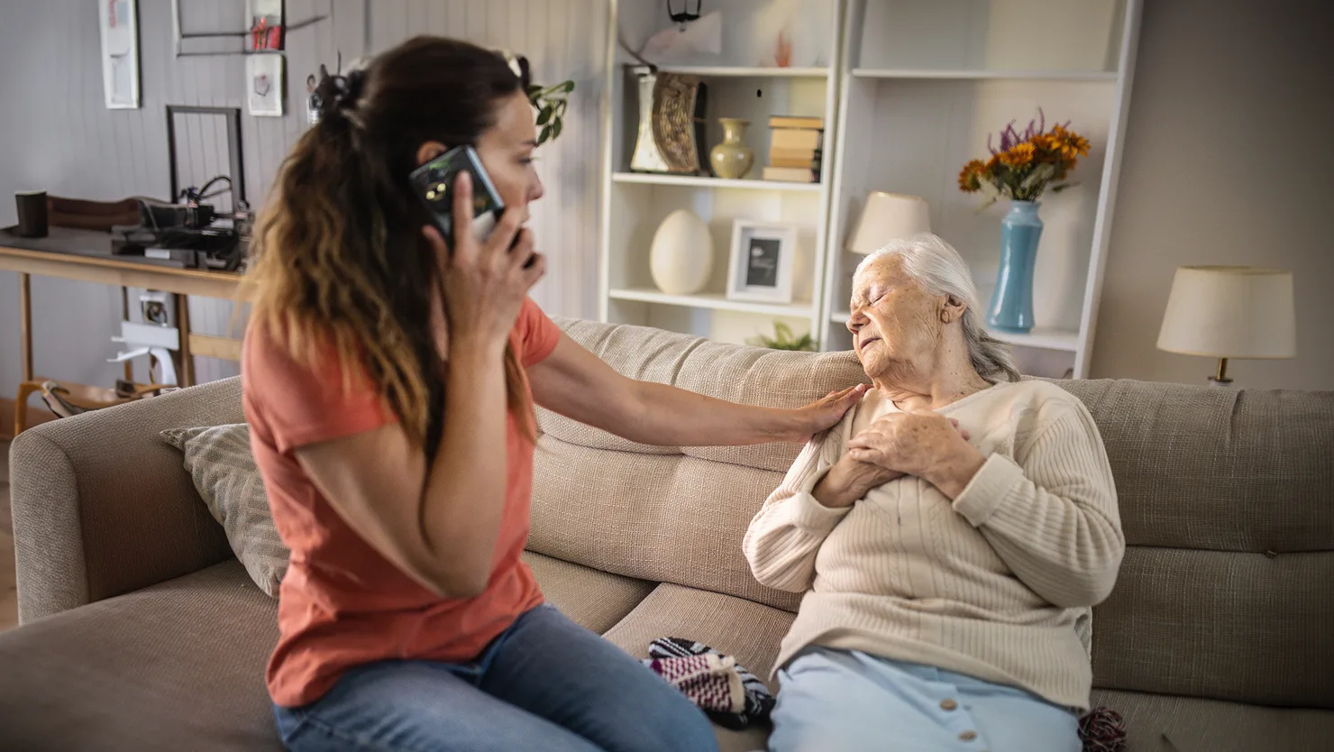 photo of a woman talking on the phone while sitting on the couch with an elderly lady laid out with her eyes closed on the couch