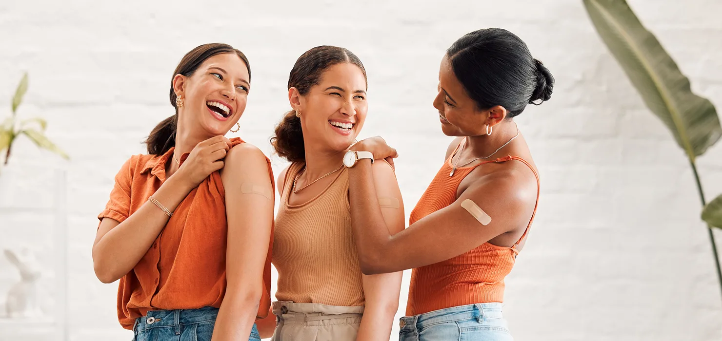 three women in their twenties smiling & laughing together as they get vaccinated