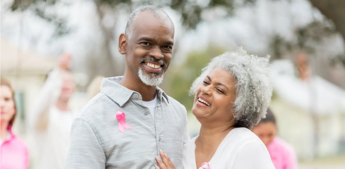 older black couple smiling & looking at the camera outside, the man is wearing a breast cancer awareness ribbon
