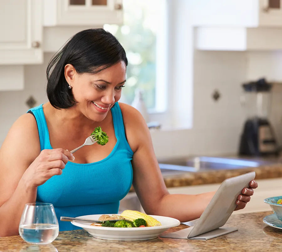image shows a woman eating a healthy meal & smiling as she reads a tablet