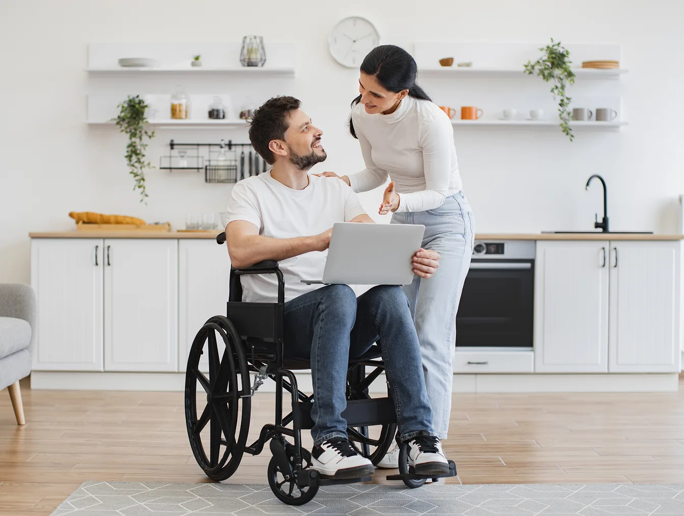 A woman standing behind a man in a wheelchair as they look at a laptop together in a kitchen