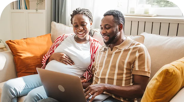 photo of a couple sitting on their couch & smiling while they look at their laptop, the woman is pregnant