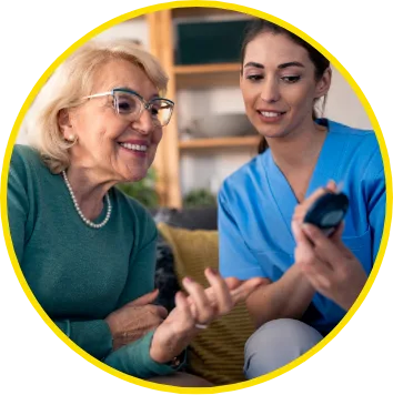 Smiling older woman sitting on a couch receiving assistance from a healthcare worker in blue scrubs, who is showing her how to use a medical device. The image is framed in a yellow circular border.